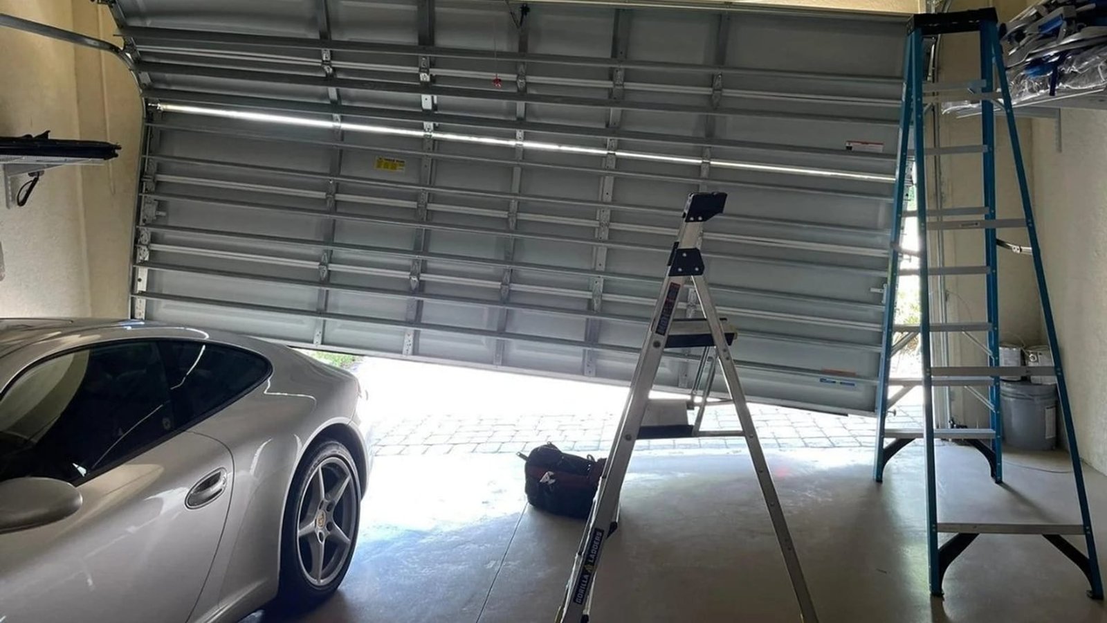Sectional garage door lifted at an angle and propped on a ladder, with a silver car in the garage