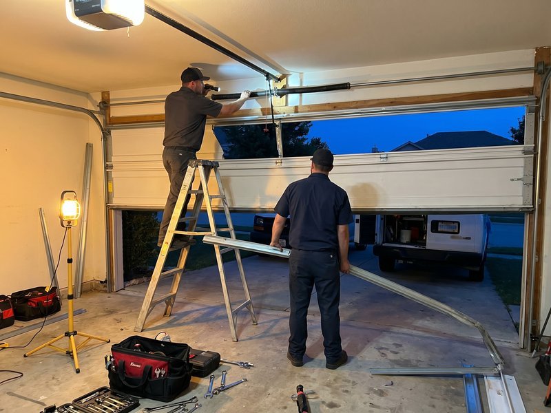 Technician on a ladder at the header of an open garage door while a second man holds a rail, a work light on the floor
