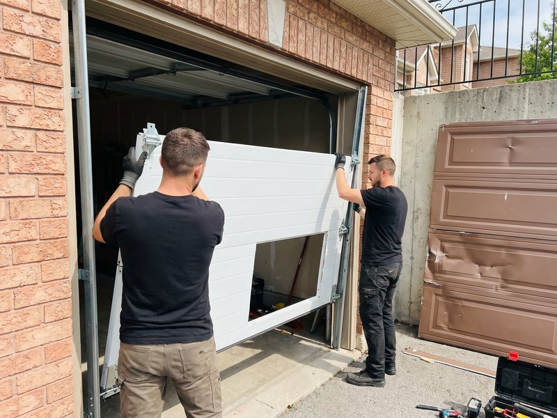 Two installers lifting a white door section into place on a brick-front garage